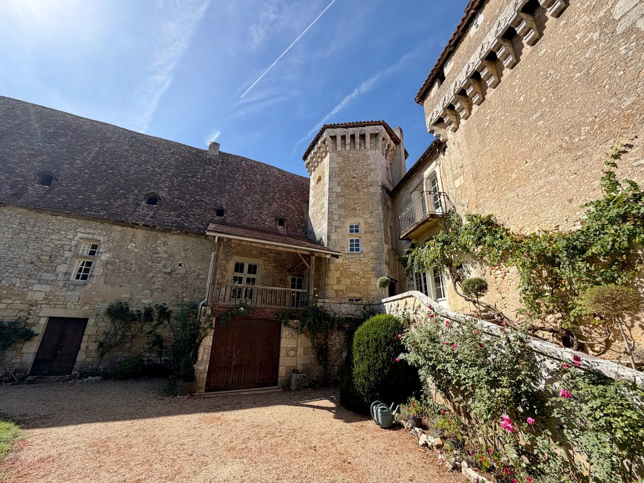 Cour gravillonnée d’un logis en pierre avec une tour carrée à mâchicoulis, un escalier bordé de rosiers roses et deux arrosoirs posés au pied des marches.