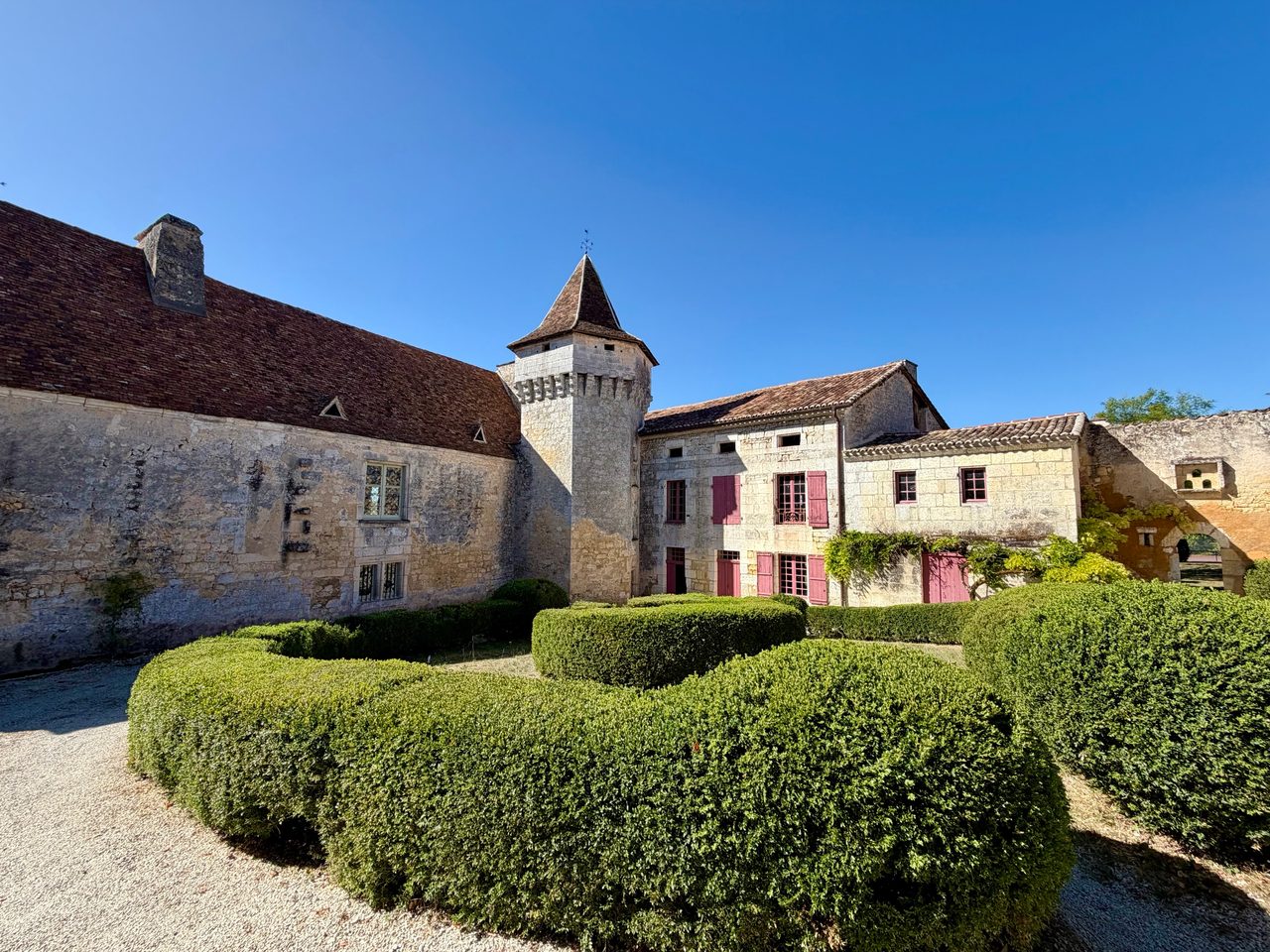 Cour d’une demeure en pierre claire avec une tour carrée à toit pointu, des volets et portes rouges, et des buis taillés en courbes au premier plan.