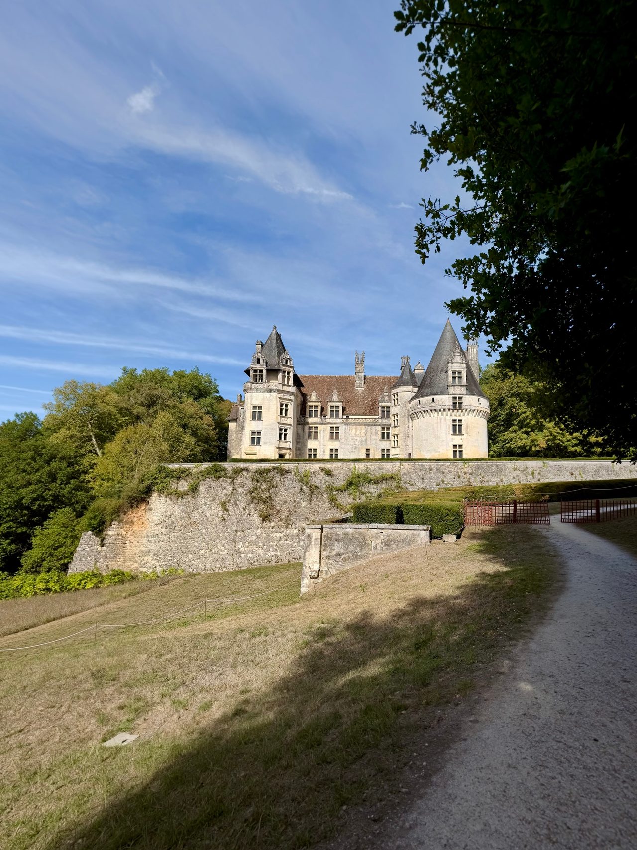 Château Renaissance blanc aux toits d’ardoise, avec une grande tour ronde, dominant un mur de soutènement et un chemin qui monte en courbe.