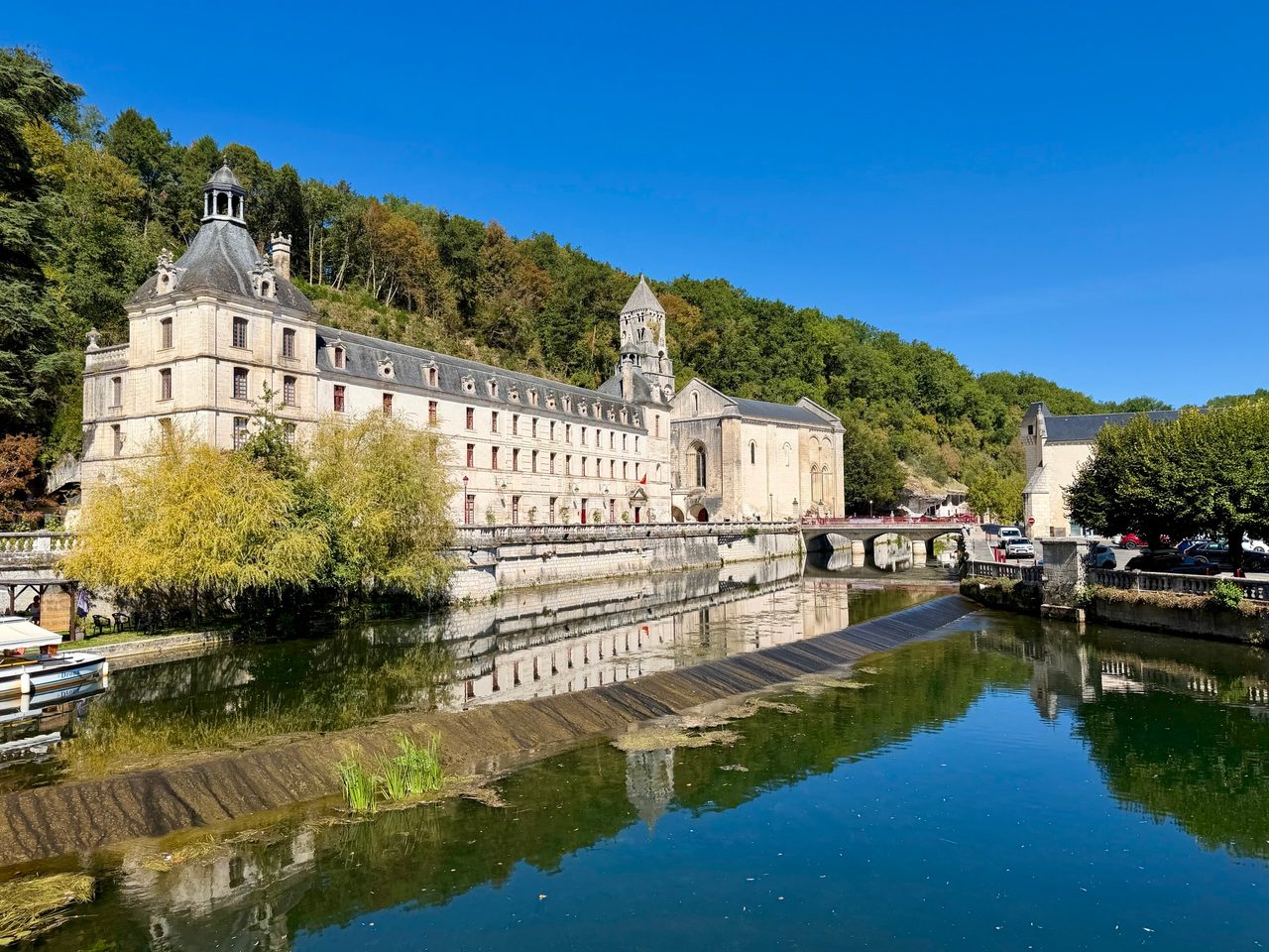 Façade longue de l’abbaye de Brantôme et son clocher, reflétés dans la Dronne au niveau d’un petit barrage.