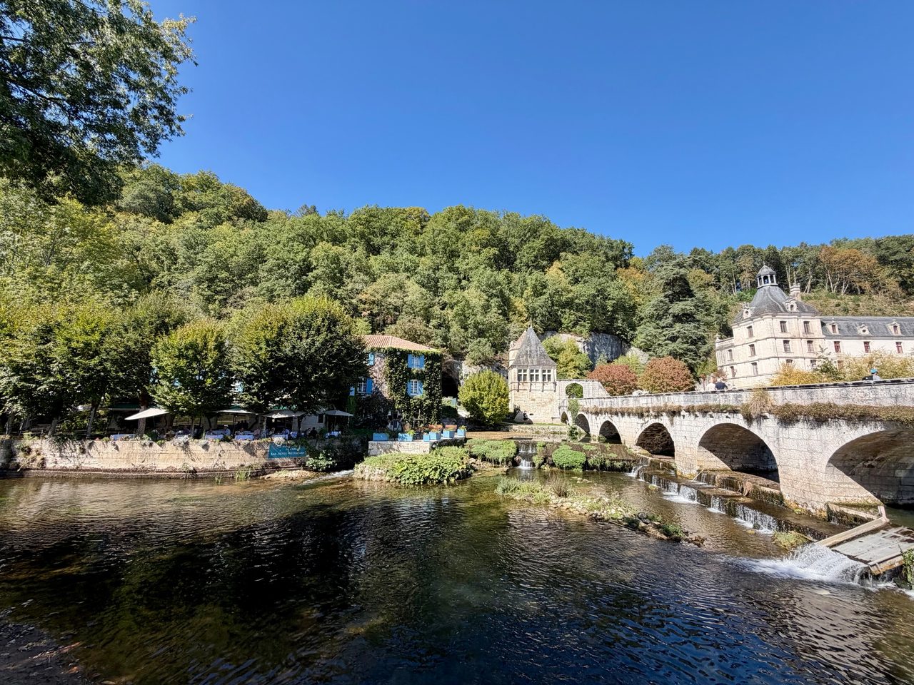 Pont de pierre à arches sur la Dronne, avec une maison couverte de lierre aux volets bleus et un pavillon de pierre au bord de l’eau.