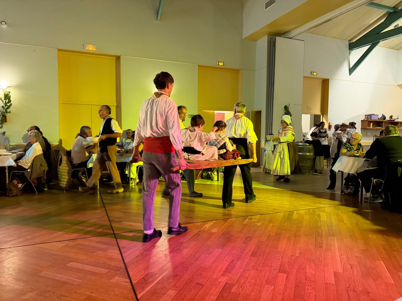 Dans une salle, deux hommes en chemise blanche et ceinture de tissu, vus de dos, portent une longue pièce de bois sur laquelle sont assis deux enfants en costume, entre des tables de convives.