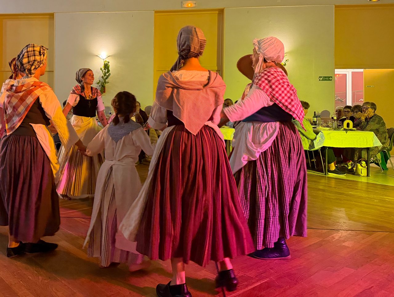 Des femmes et une fillette en costume traditionnel, jupes longues rayées, fichus à carreaux et coiffes, dansent en ronde en se tenant la main sur un parquet, sous des éclairages roses et jaunes.